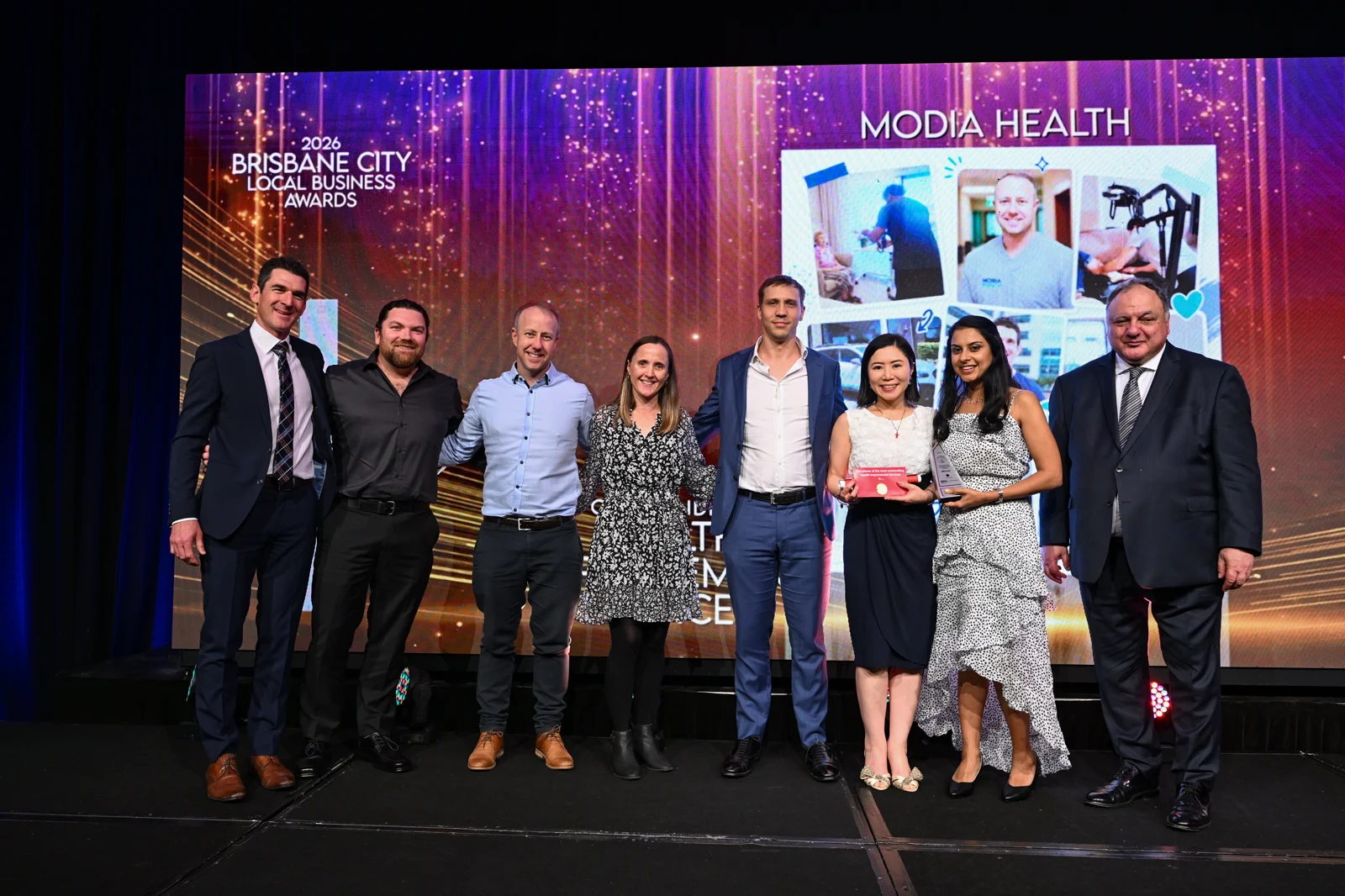 The Modia Health group on the awards stage beneath a screen displaying the company’s name.